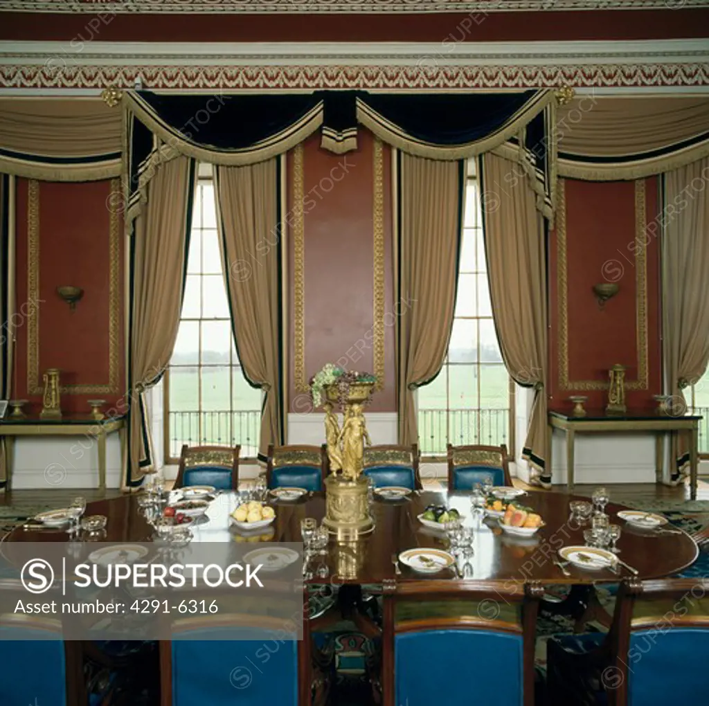 Ornate swagged curtains in large country house dining room with blue chairs and place settings on the table