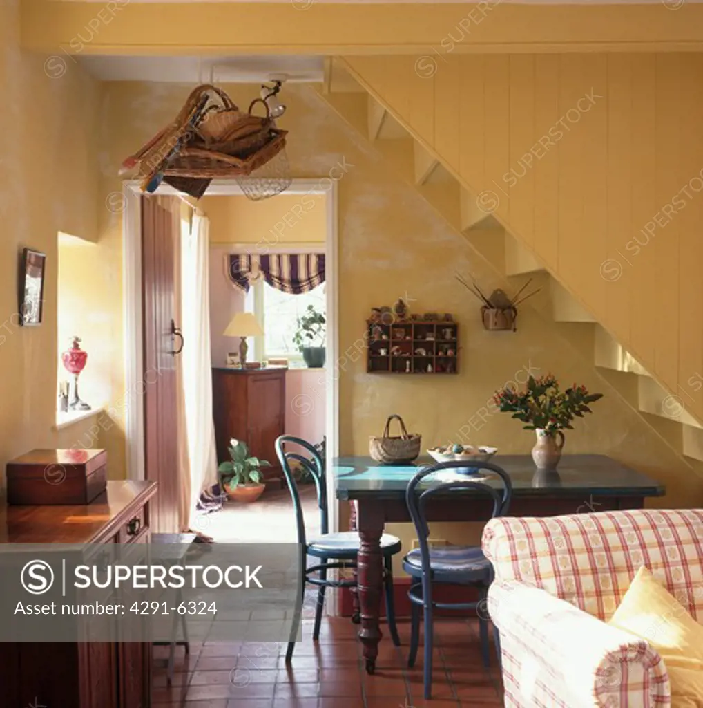 Bentwood chairs and wooden table below staircase in yellow cottage kitchen with terracotta tiled floor and red check sofa