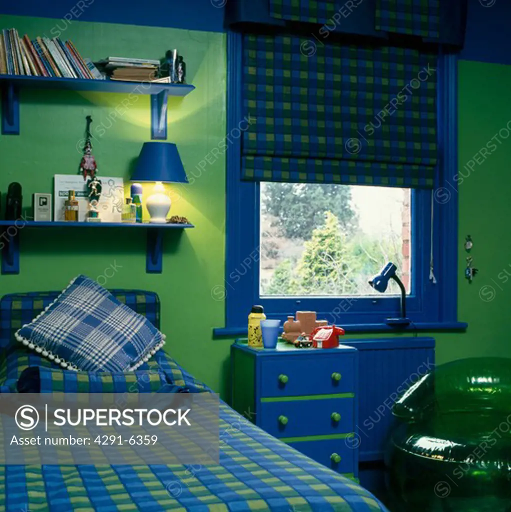 Blue and green checked bedlinen on bed below blue shelves in child's green bedroom with blue paintwork and blue and green checked blind