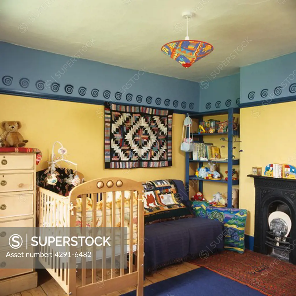 Pine cot and patchwork wall-hanging below stencilled pattern on blue and yellow wall in nursery bedroom