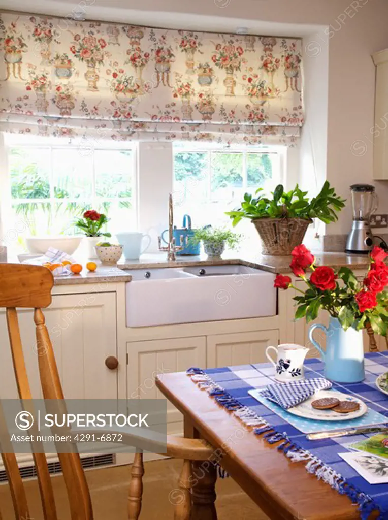 Floral blind on window above Belfast sink in cream country kitchen with blue checked cloth on wooden table