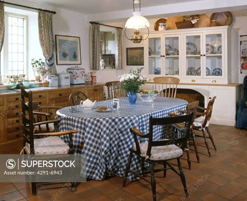 Antique chairs around table with blue and white checked PVC cloth in country kitchen with white dresser