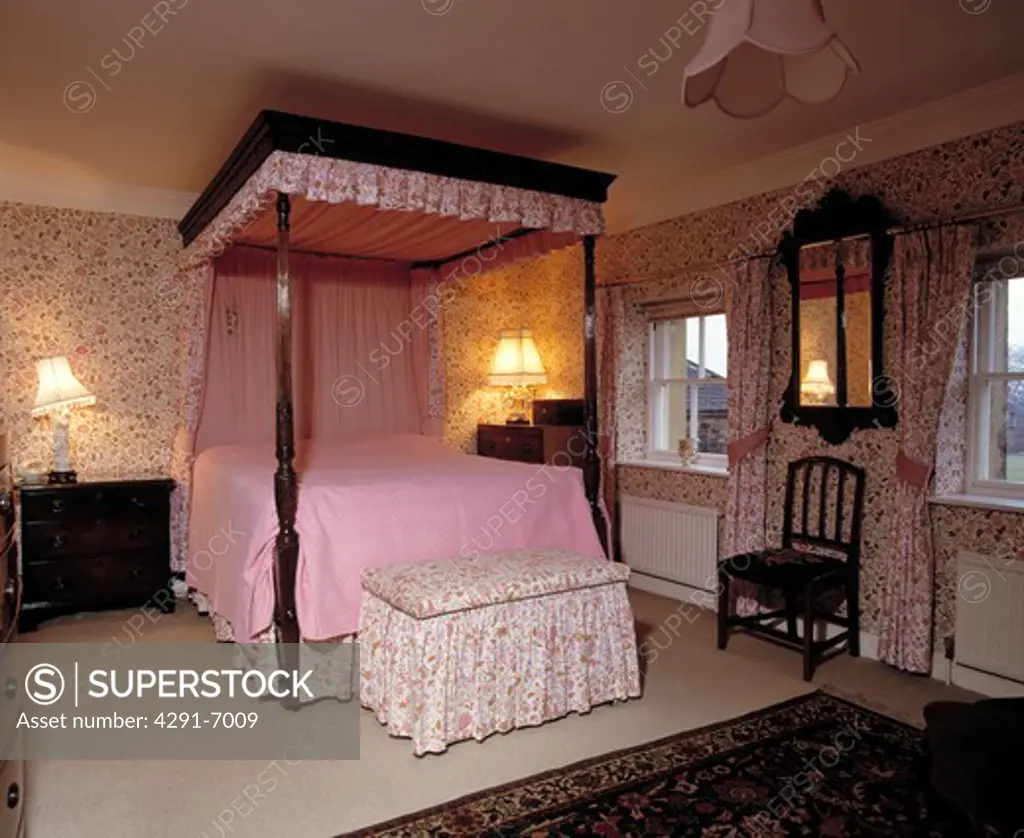 Pink bedcover on four-poster bed in country bedroom with floral wallpaper and floral loose-cover on ottoman