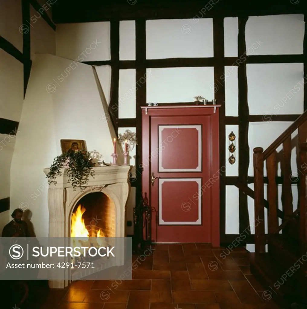 Stone mantelpiece on corner fireplace with lighted fire in country hall with black beams and red door