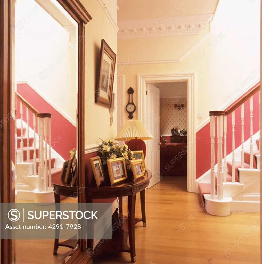 Large mirror and photographs on table in hall with wooden flooring and red dado on staircase