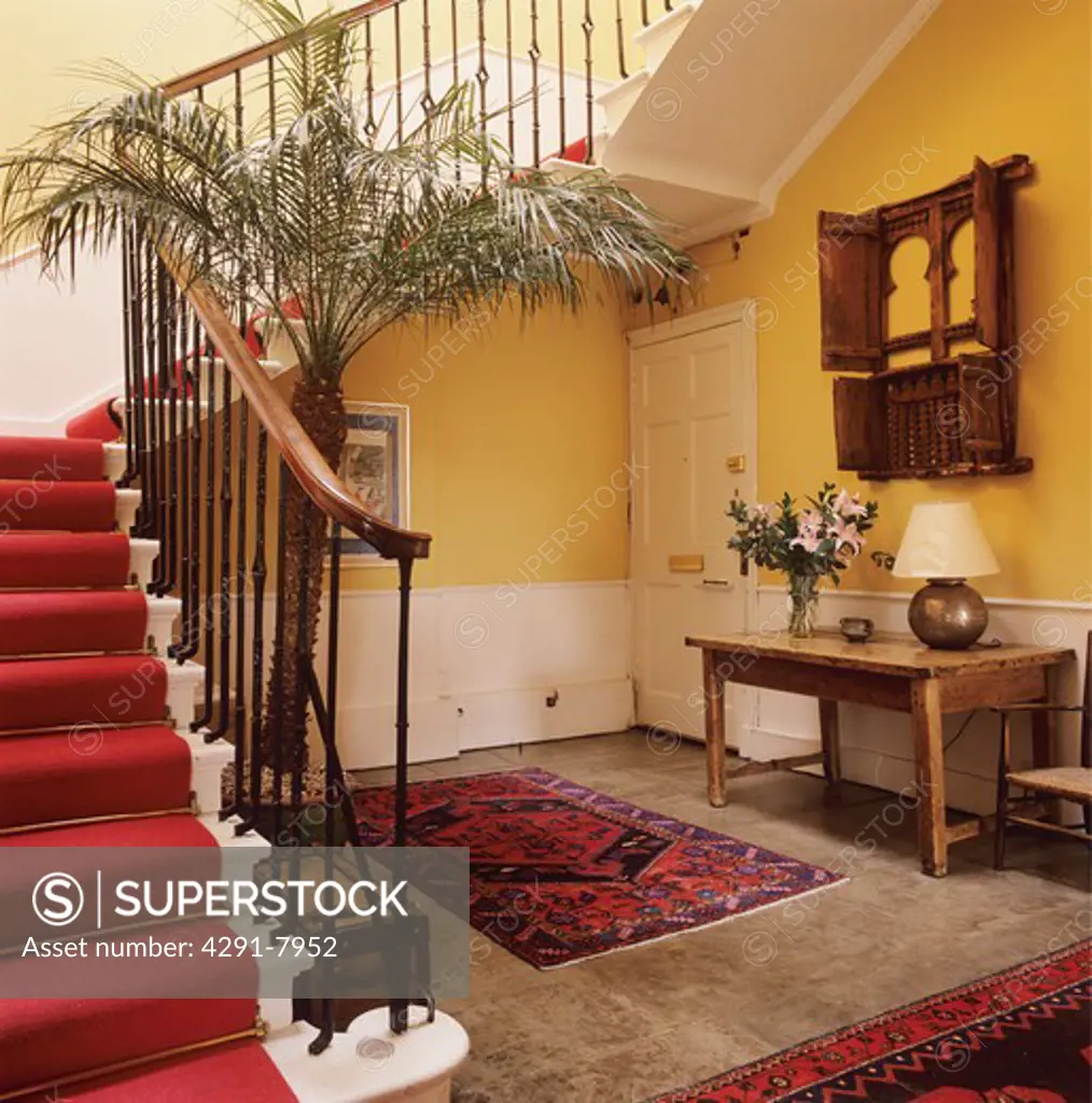 Red carpet on staircase beside palm houseplant in yellow hall with rugs on stone-flagged floor