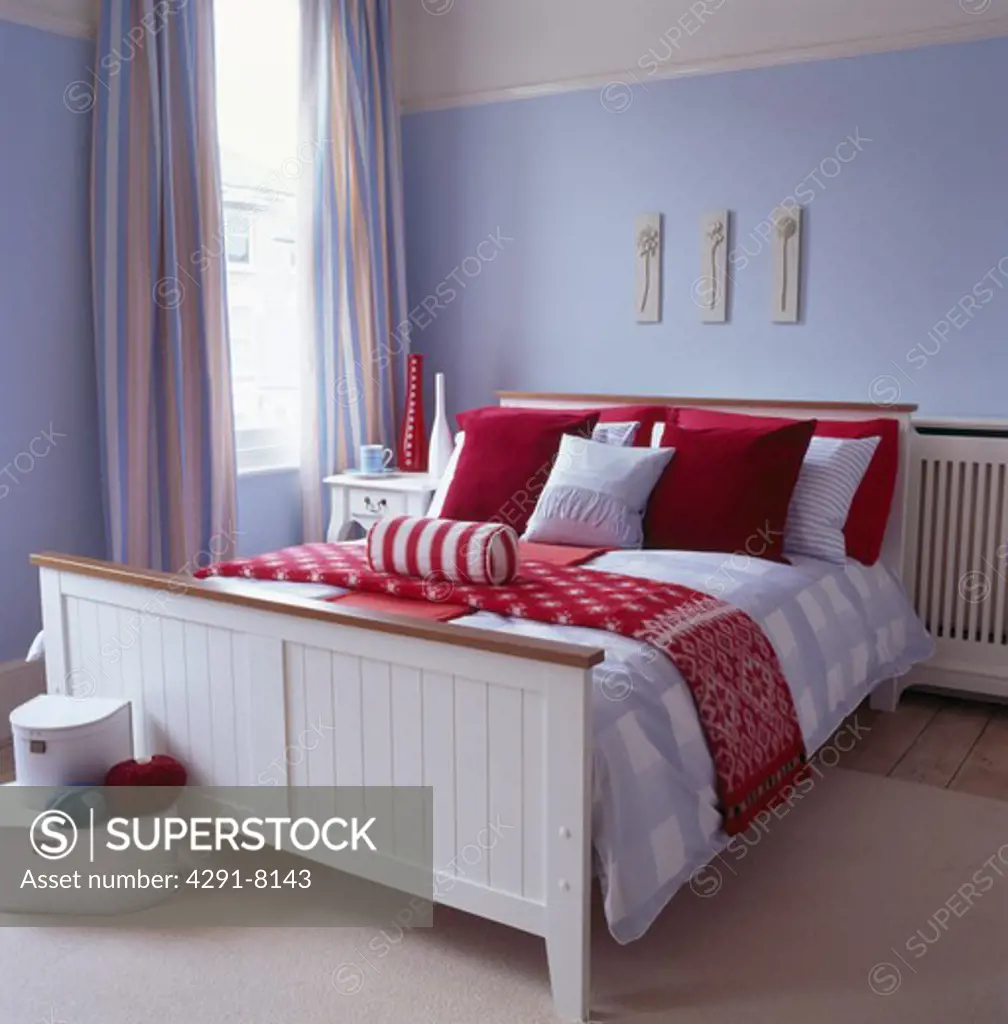 Red and white cushions and bedlinen on white wooden bed in pastel blue bedroom