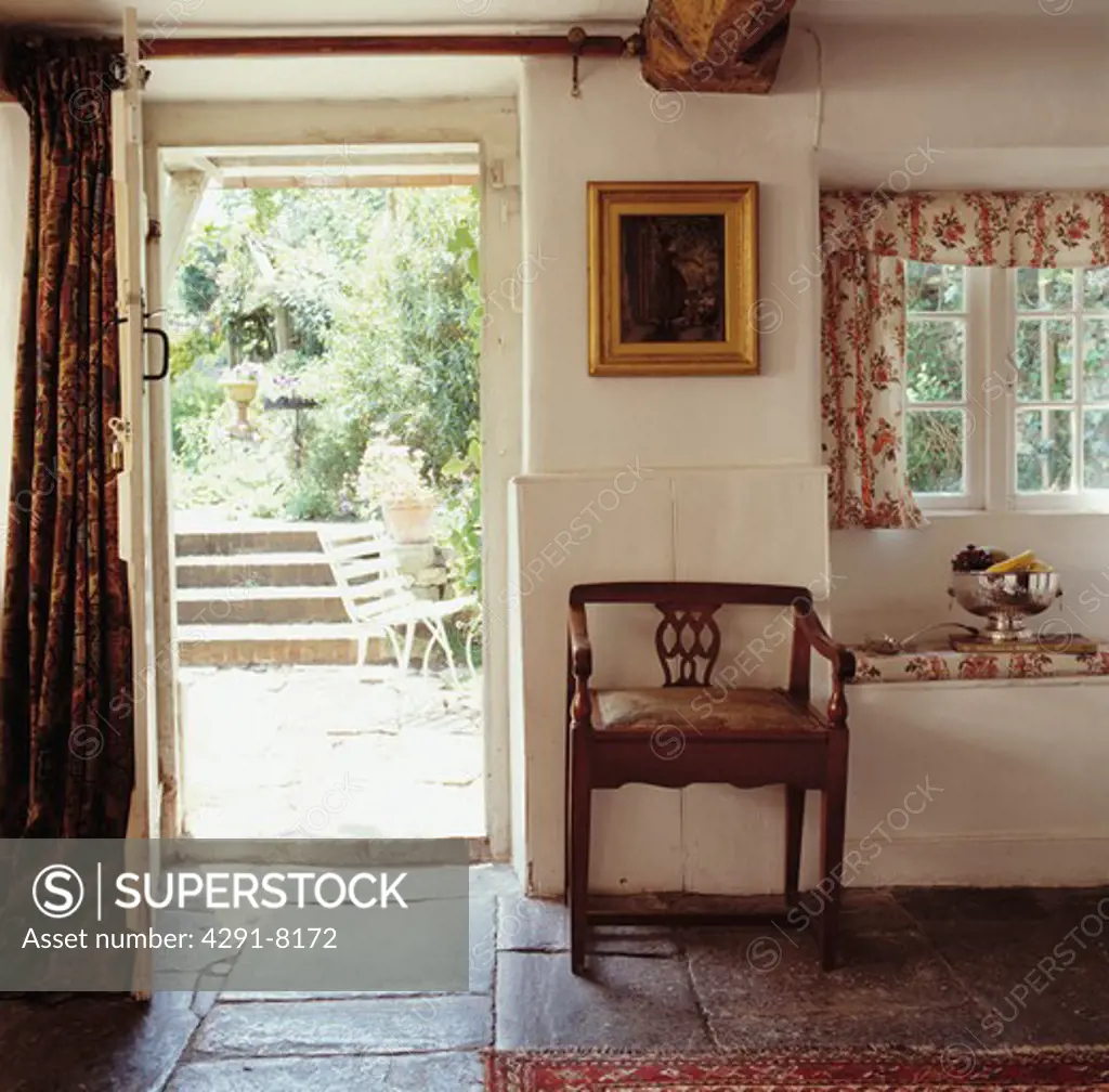 Cottage hall with stone-flagged floor and antique chair beside open door with view of the garden