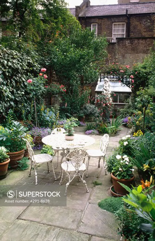 Plants in pots & white metal table and chairs on stone paved patio in town garden with statue and greenhouse