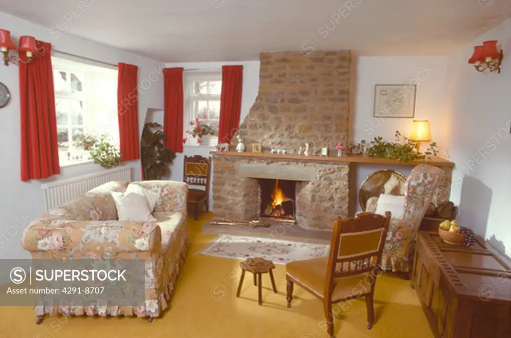 Floral loosecover on sofa in front of stone fireplace with lighted fire in seventies cottage living room