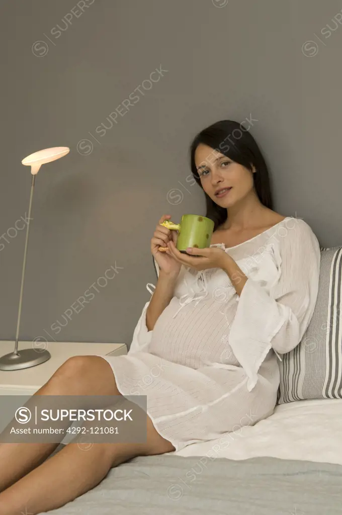 Pregnant woman sitting on bed with mug