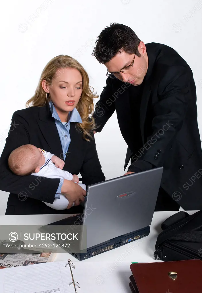 Professional woman in office, holding baby looking at laptop with male colleague