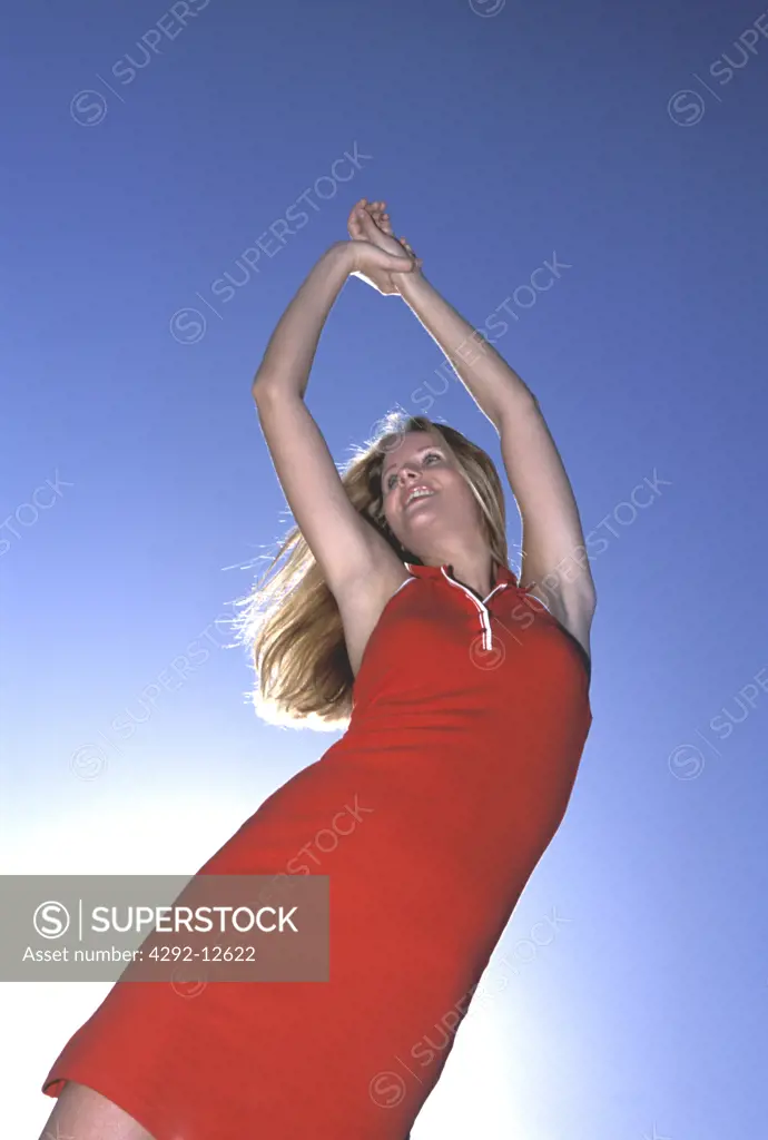 Young red dressed woman standing and stretching arms against blue sky