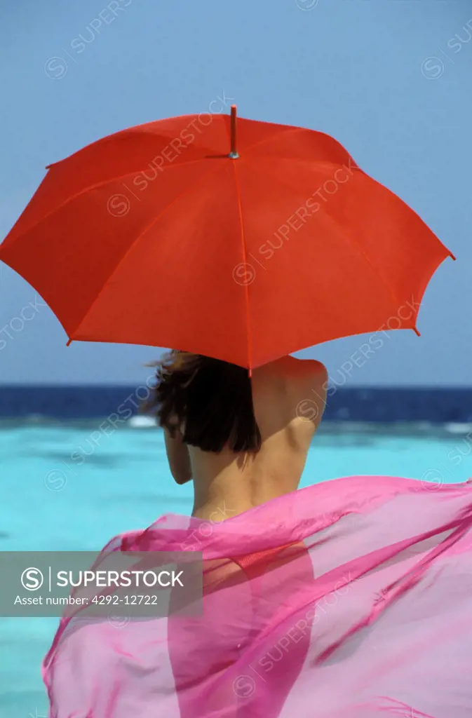 toplesee Woman standing on seashore holding an umbrella, looking at the horizon, with a flying pareu