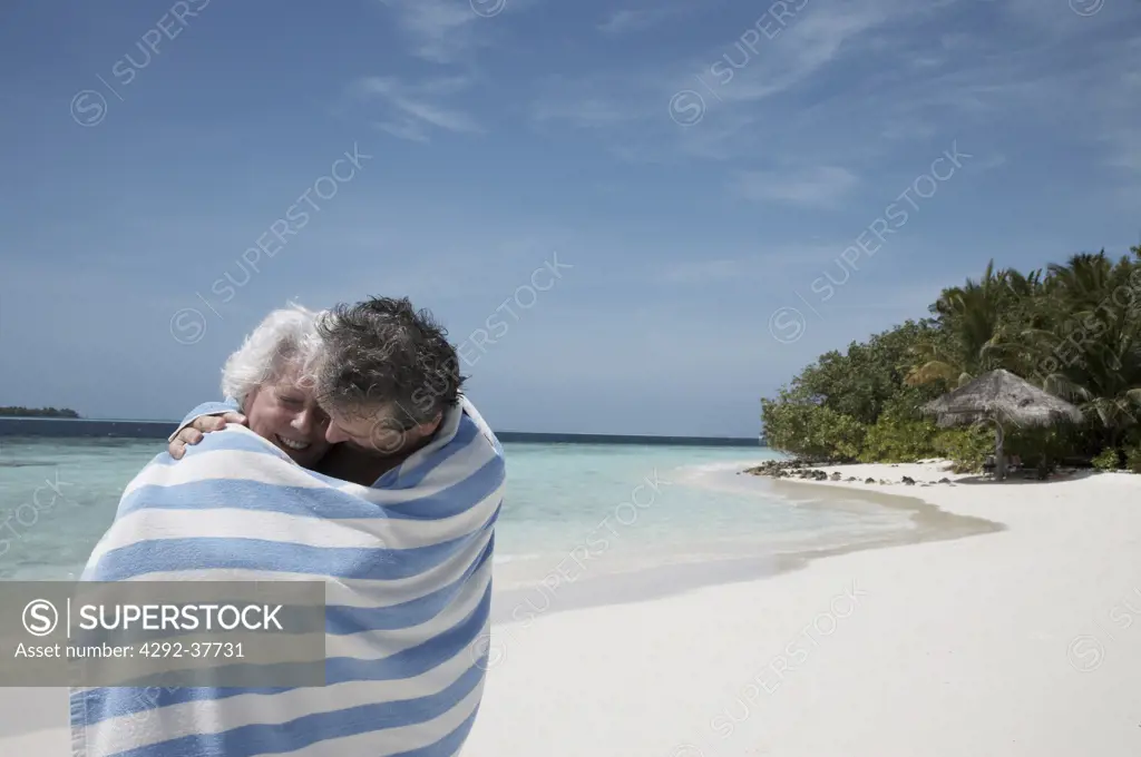 Maldives, Ari Atoll, senior couple embracing wrapped in a beach towel