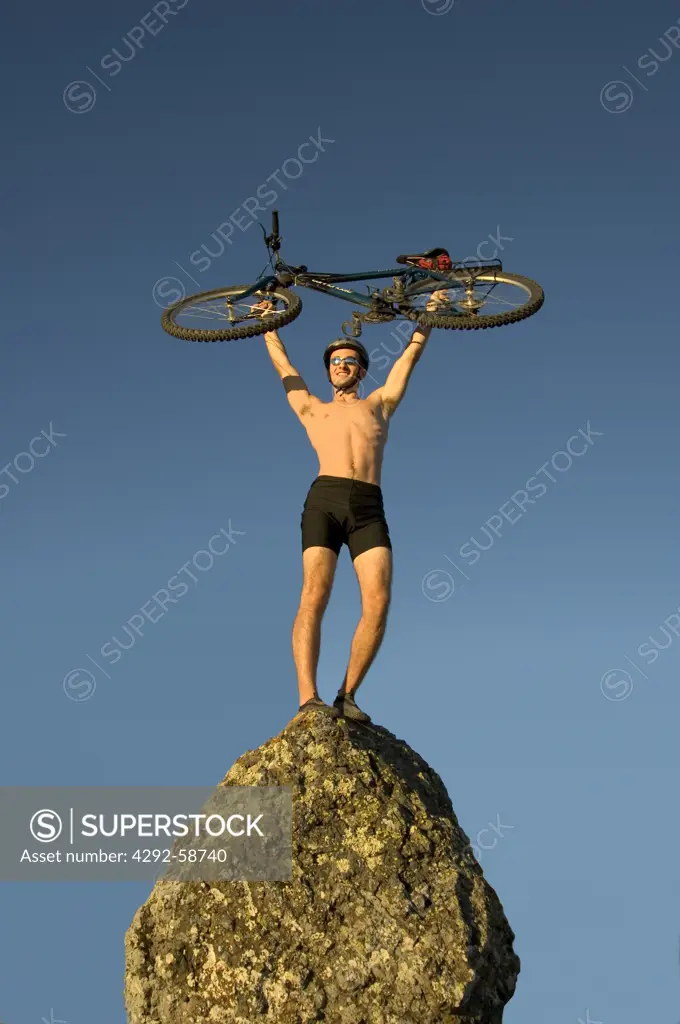 Man standing on mountain top lifting bicycleoverhead celebrating victory