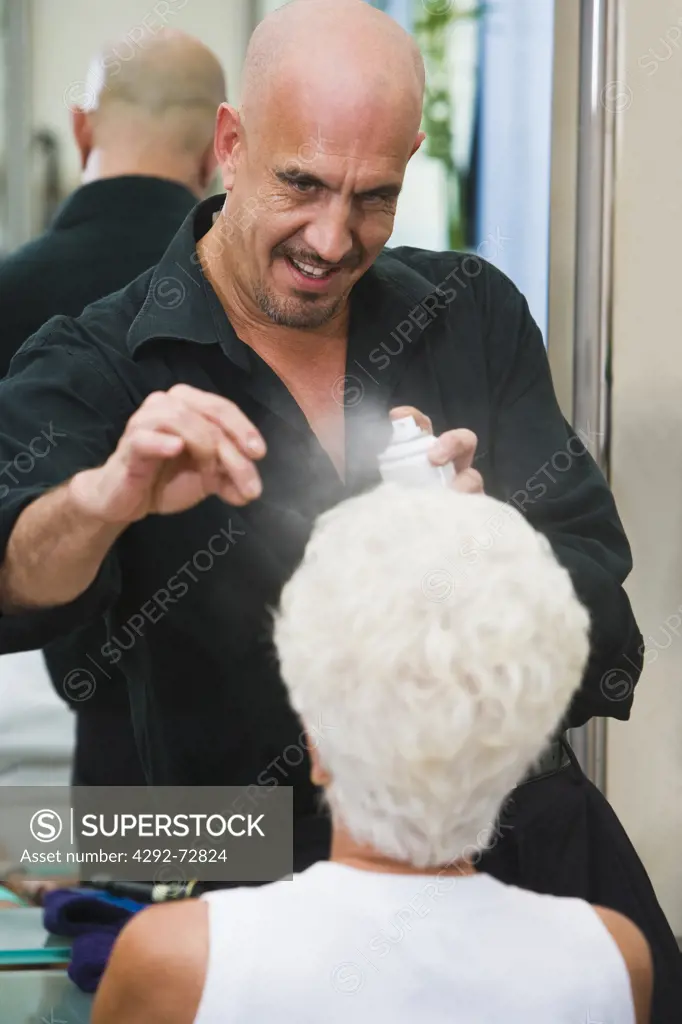 Male hairdresser spraying hair to older woman at beauty salon