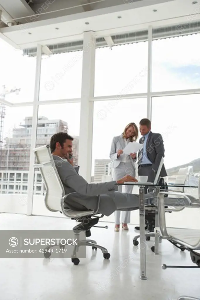 Businessman in office with legs on chair with businesspeople talking in the background