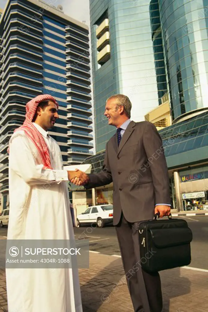 An Arab and a businessman shaking hands near a commercial complex.