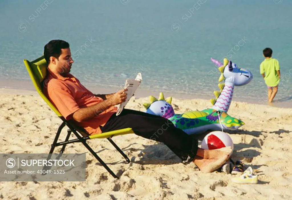 A man reading newspaper as a boy stands near the seashore.