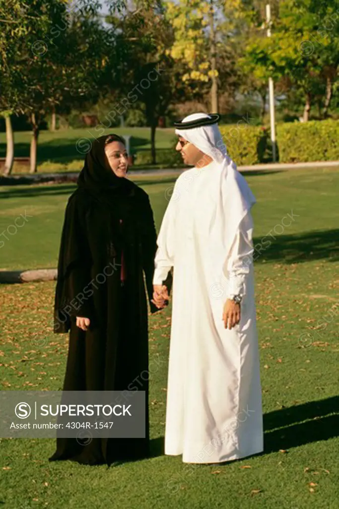 An Arab couple smiling at each other as they hold hands in the park.
