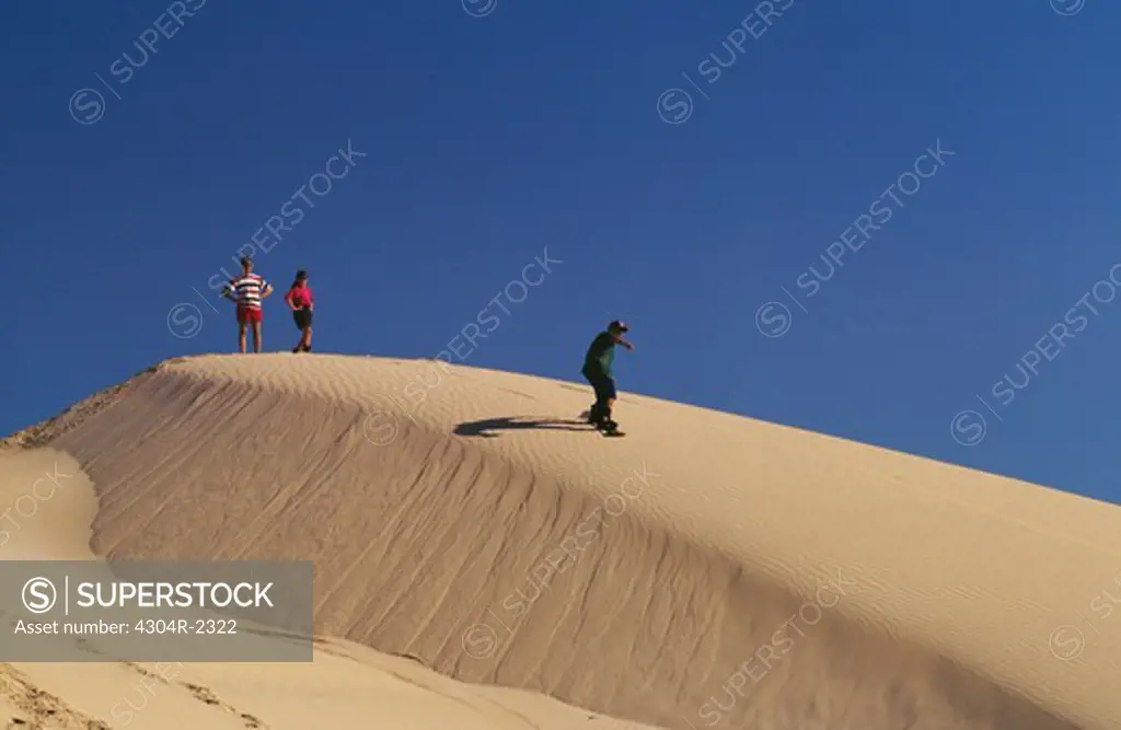 A man slides on the sand dune with a snowboard as a couple look at him.