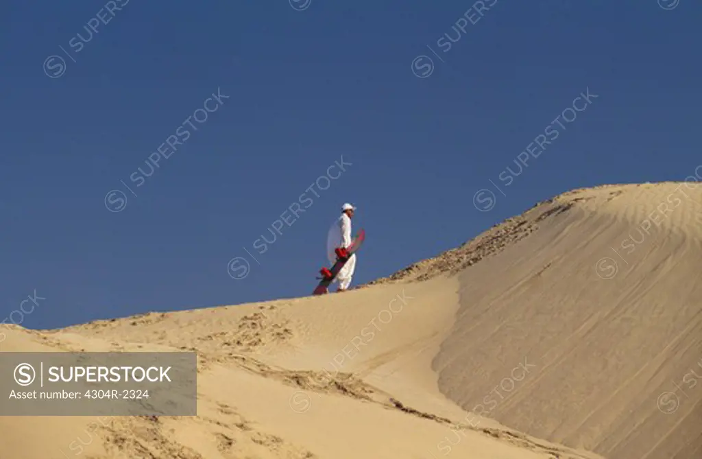 A mature man walks on the sand dune carrying a snowboard in his hand.