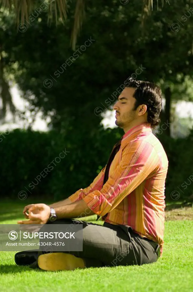 Young businessman sitting on green grass & doing yoga