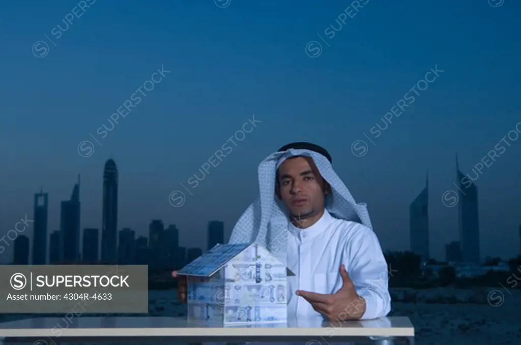 Young Arab man sitting at a desk with Dubai City in the background holding a house made of money