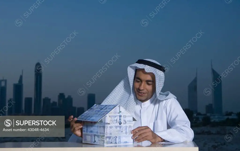Young Arab man sitting at a desk with Dubai City in the background holding a house made of money