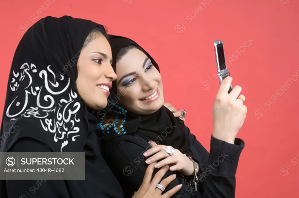 Two Arab Ladies standing cheek to cheek with cell phone