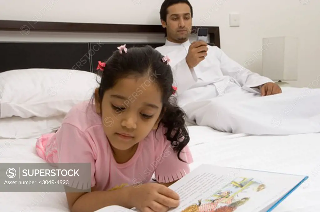 Daughter reading on the bed while father is using his cell phone