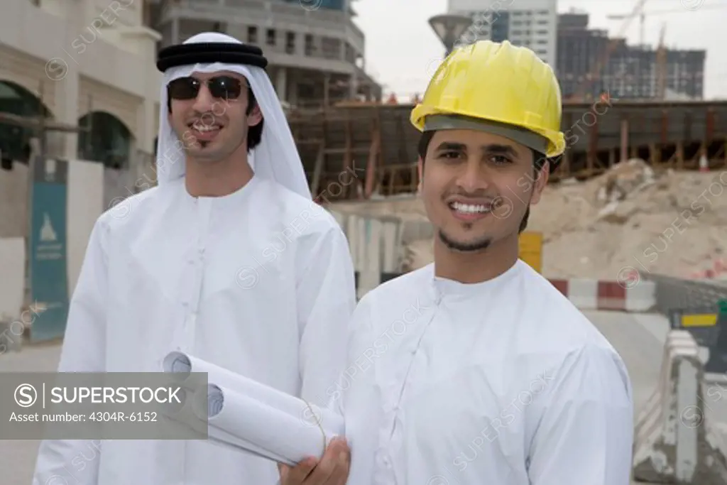 Businessmen holding blueprints with buildings in background, smiling, portrait
