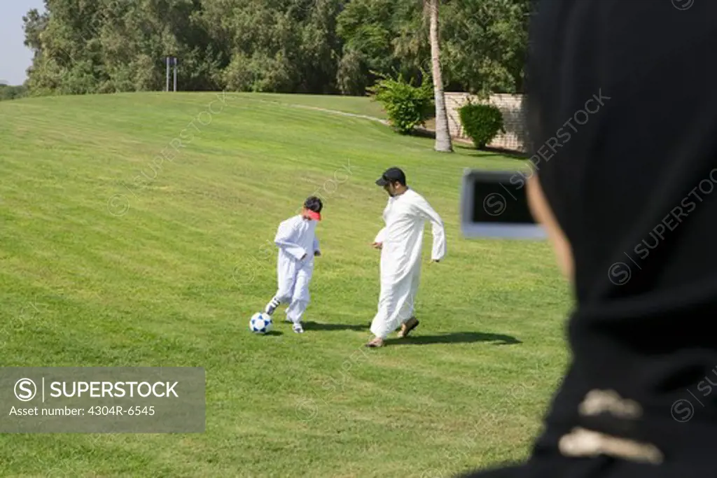 Father and son playing football at park while woman taking photograph