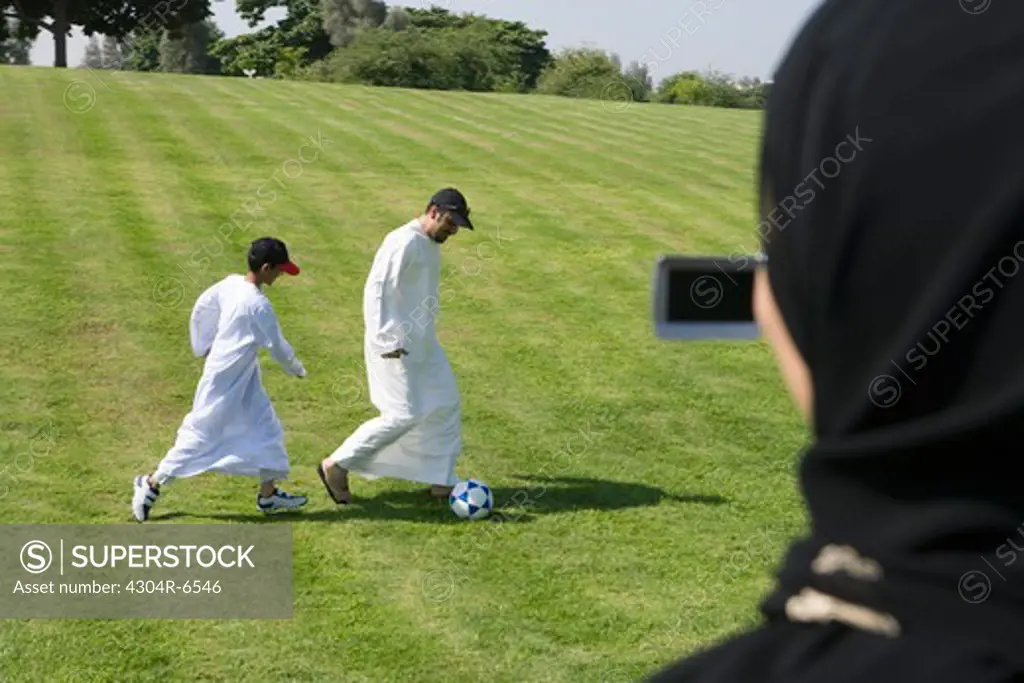 Father and son playing football at park while woman taking photograph