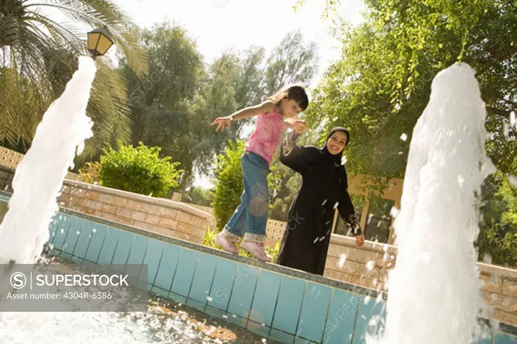 Mother with daughter standing by fountain at park, smiling