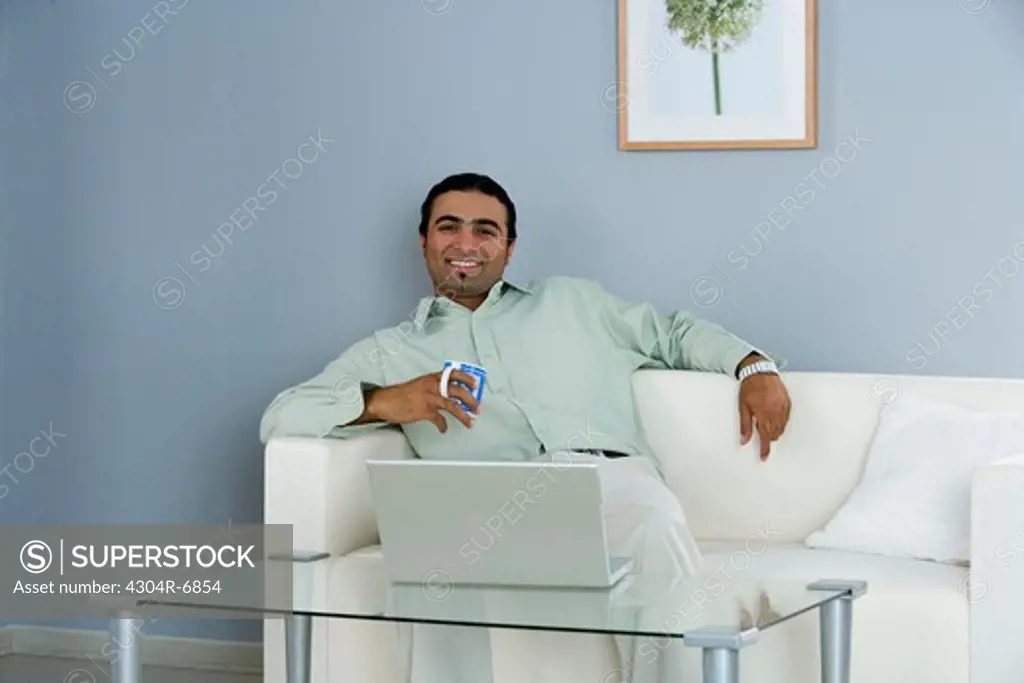 Man sitting on sofa and holding cup, laptop on the table