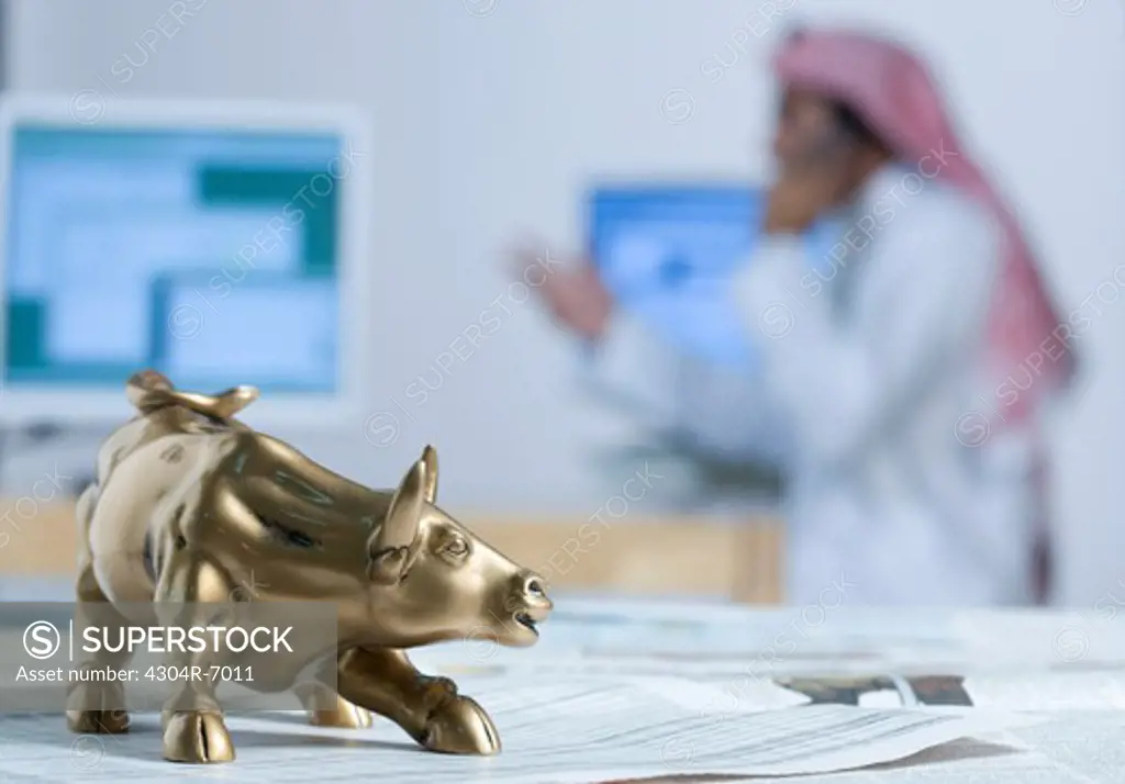 Bull figurine on newspaper, background computers and Arab man, close-up