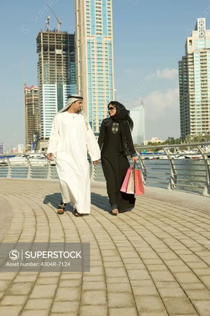 Arab couple holding hands while walking, woman with shopping bags, at Dubai Marina, UAE