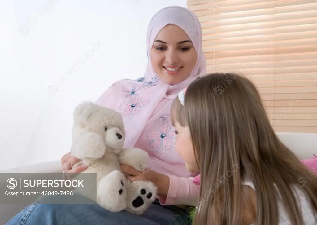 Mother and daughter sitting on sofa, playing with teddy bear