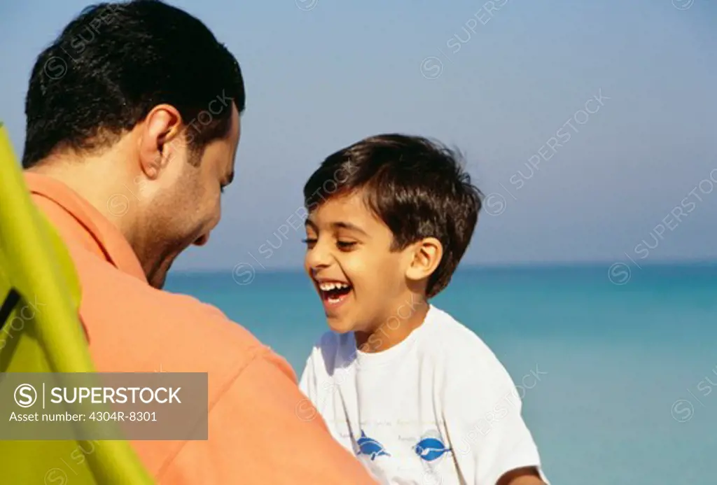 A father and son heartily smile at each other while seated on the chair as they enjoy at the beach.
