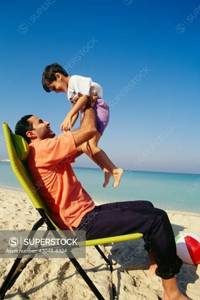 A father holds his son while seated on the chair as they enjoy at the beach.