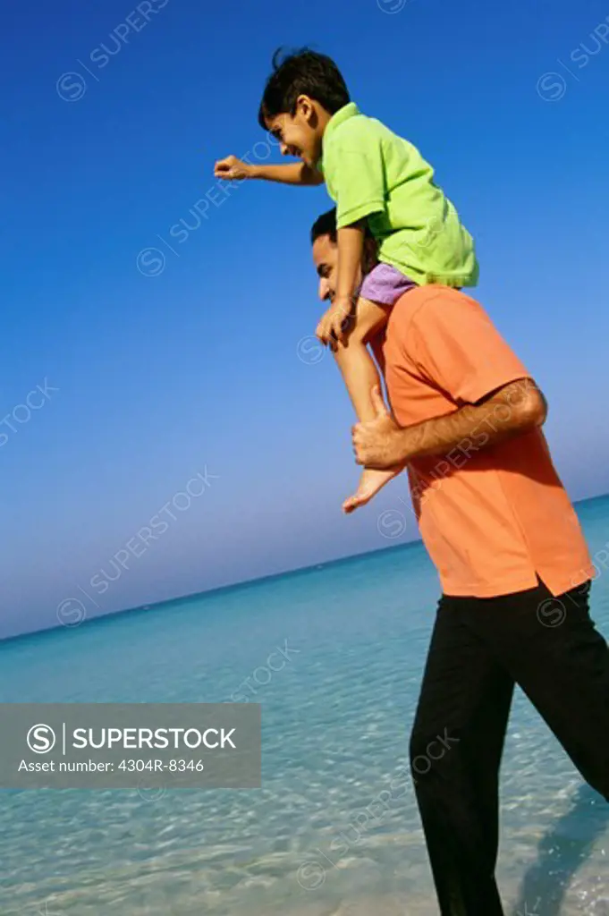 A father carries his son on his shoulders as he walks on the seashore.