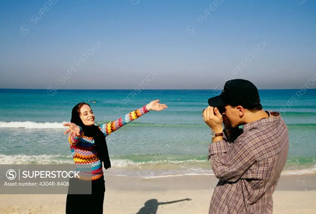 A man takes a photograph of the woman on the beach.