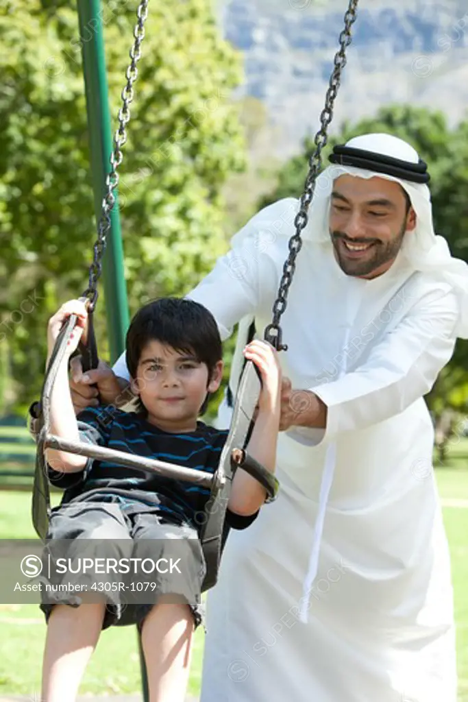 Boy sitting on swing being pushed by his arab father, smiling.