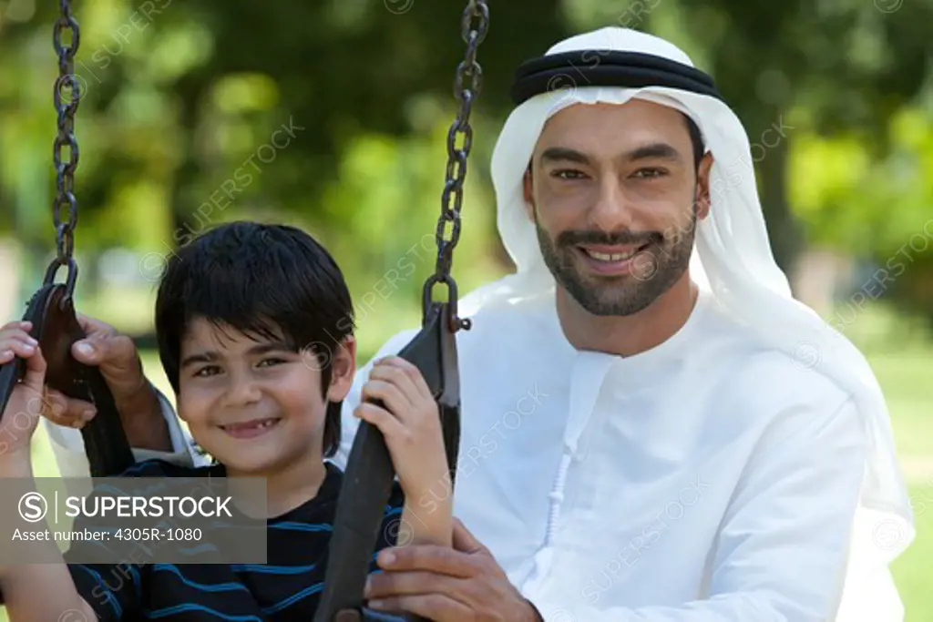 Portrait of an arab father and son at the park, boy sitting on swing.