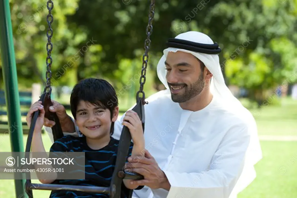 Arab father and son playing at the park, boy sitting on swing.