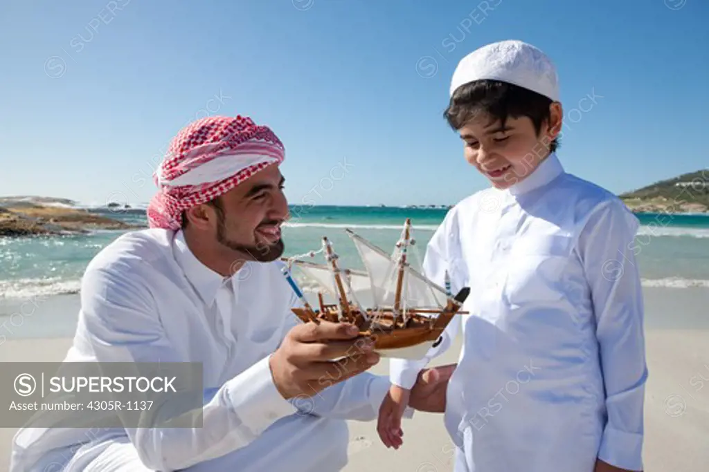 Arab father and son with toy boat on beach.