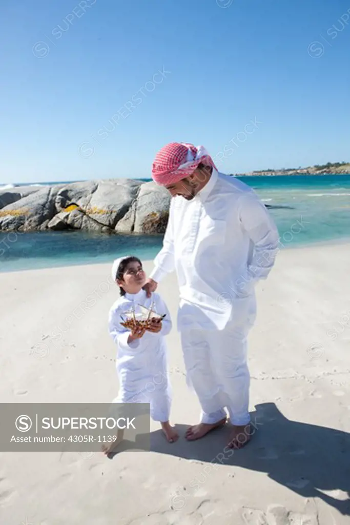 Arab father and son with toy boat walking on beach.