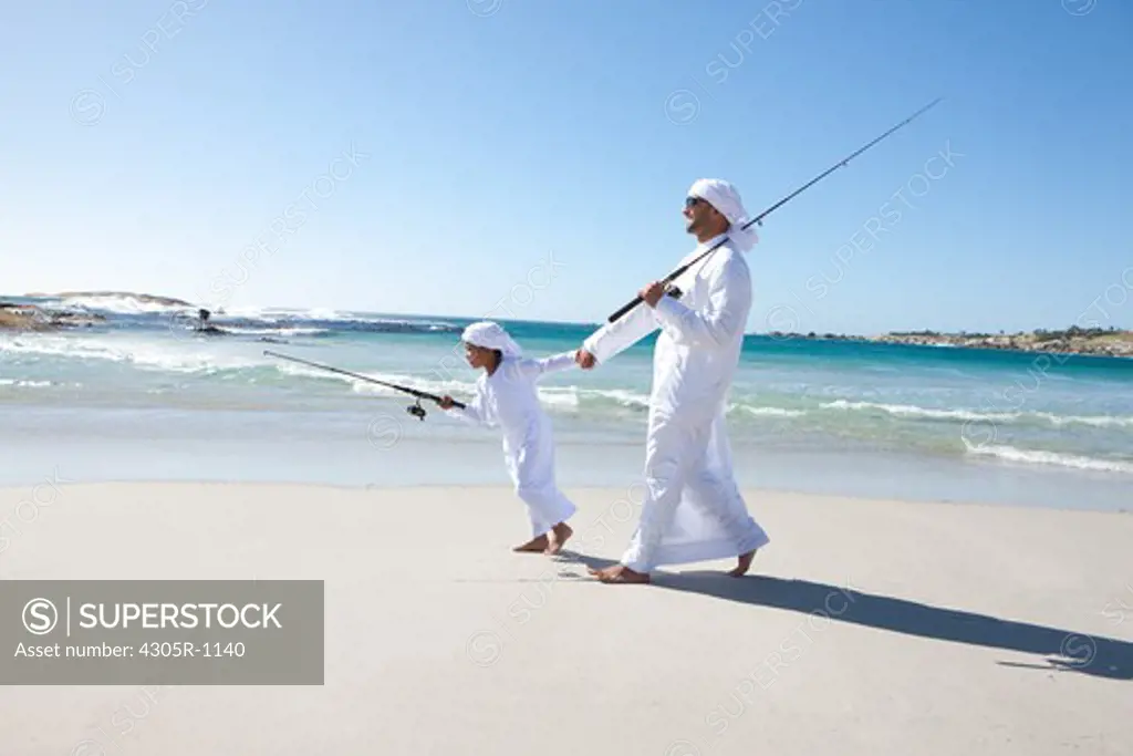 Arab father and son with fishing rod walking on beach.